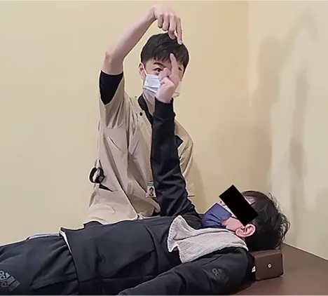 仰向けの利用者が天井に向けて手を伸ばすトレーニングをしている。